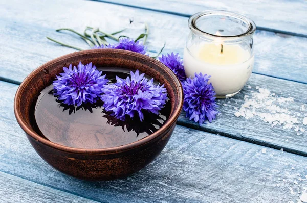 Fiori di mais blu si trovano in acqua in una ciotola vicino a una candela accesa sullo sfondo delle vecchie tavole blu . — Foto Stock