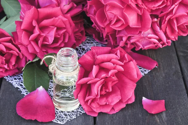 Botella de vidrio con aceite esencial de rosa y rosas rojas sobre un fondo de madera oscura . — Foto de Stock