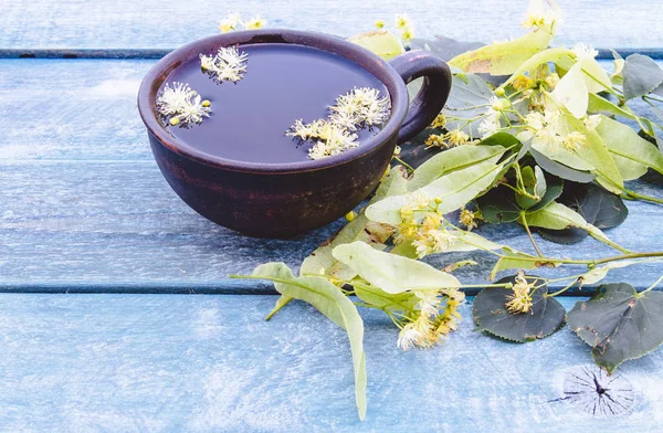 Una tazza di tè di tiglio vicino ai fiori di tiglio sullo sfondo delle vecchie tavole blu. fiori di tiglio e una tazza di tè . — Foto Stock