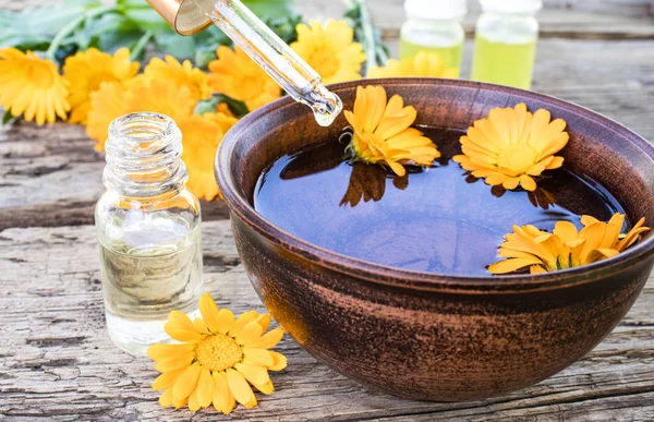 Calendula essential oil near the yellow flowers of calendula on a wooden background. Extract of calendula tincture in a bowl. — Stock Photo, Image