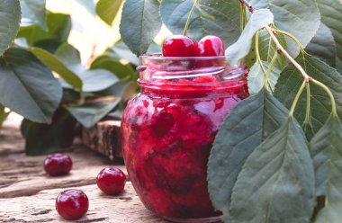 Ahşap tahta bir masada yeşil yaprakları bir arka planda bir cam kavanoz kiraz reçel.