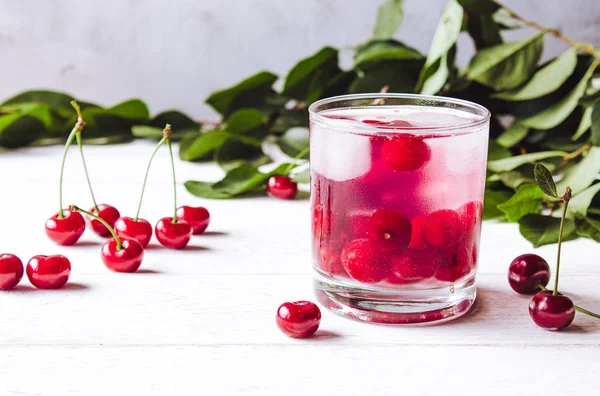 Red cocktail with cherry and ice on a white wooden background. Fresh summer cocktail with cherries and ice cubes.