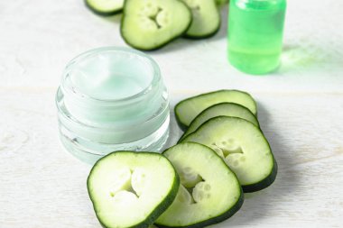 Beyaz ahşap arka planda taze salatalık dilimleri yanında bir cam kavanoz yüz kremi.