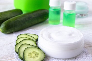 Beyaz ahşap arka planda taze salatalık dilimlerinin yanında salatalık özü ile beyaz bir kavanozda yüz kremi.