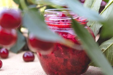 Ahşap tahta bir masada yeşil yaprakları bir arka planda bir cam kavanoz kiraz reçel.