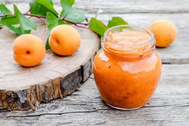 Ahşap bir yüzey üzerinde olgun kayısı ve yeşil yapraklarıyanında cam bir kavanozda kayısı reçeli.