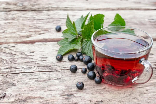 Yeşil yaprakları ve kuş üzümü çilek yanında ahşap bir masada siyah kuş üzümü ile bir fincan çay.