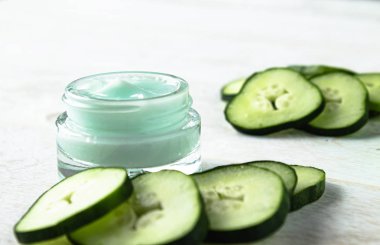 Beyaz ahşap arka planda taze salatalık dilimleri yanında bir cam kavanoz yüz kremi.