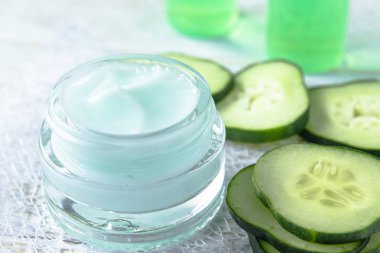 Beyaz ahşap arka planda taze salatalık dilimleri yanında bir cam kavanoz yüz kremi.