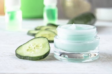 Beyaz ahşap arka planda taze salatalık dilimleri yanında bir cam kavanoz yüz kremi.