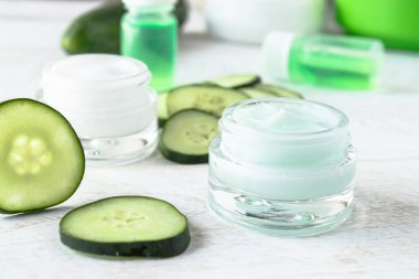 Beyaz ahşap arka planda taze salatalık dilimleri yanında salatalık özü ile bir cam kavanoz yüz kremi.