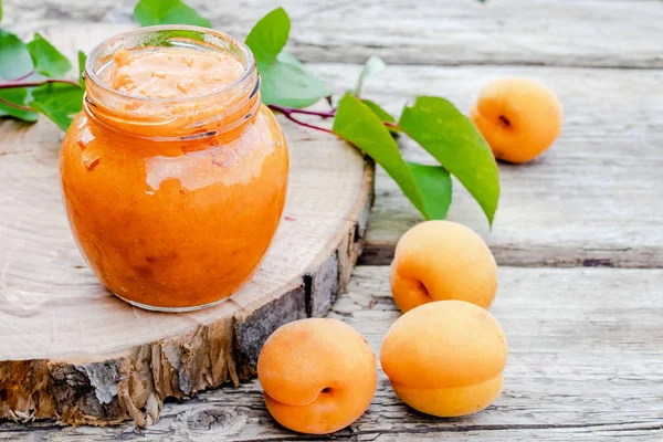 Confiture d'abricot dans un bocal en verre à côté d'un abricot mûr et de feuilles vertes sur une surface en bois . — Photo