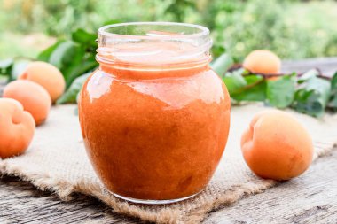 Olgun kayısı ve yeşil yaprakların yanında ahşap bir yüzeyüzerinde ev yapımı kayısı reçeli ile cam bir kavanoz.