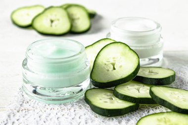 Beyaz ahşap arka planda taze salatalık dilimleri yanında bir cam kavanoz yüz kremi.