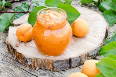 Ahşap bir yüzey üzerinde olgun kayısı ve yeşil yapraklarıyanında cam bir kavanozda kayısı reçeli.