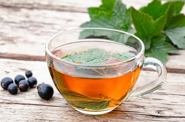 Herbal tea from currant leaves in a glass transparent cup on a wooden table near the green leaves and currant berries.