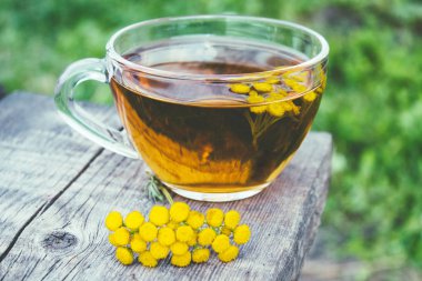 Doğanın arka planına karşı ahşap bir masanın yüzeyinde bir kupa ve sarı tansy çiçekler bitkisel çay veya tansi tentür. Şifa otlar.