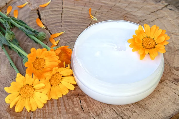 Ein Glas weißer Kosmetikcreme für die Körperpflege. frische orange Ringelblumen auf hölzernem Hintergrund. — Stockfoto