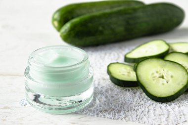 Beyaz ahşap arka planda taze salatalık dilimleri yanında bir cam kavanoz yüz kremi.