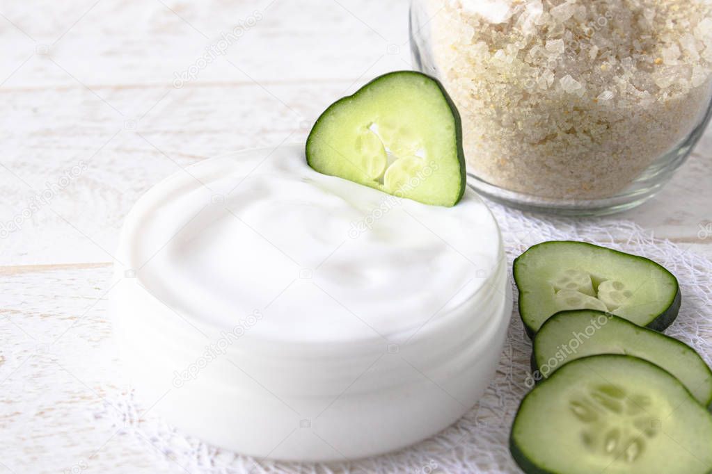 Face cream in a white jar with slices of fresh cucumbers on a white wooden background.