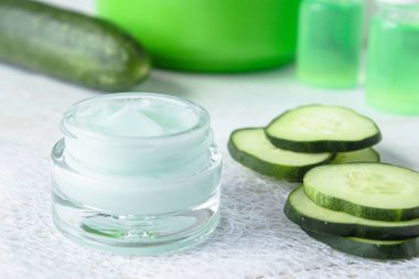 Beyaz ahşap arka planda taze salatalık dilimleri yanında bir cam kavanoz yüz kremi.