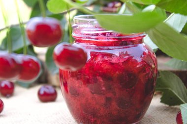 Ahşap tahta bir masada yeşil yaprakları bir arka planda bir cam kavanoz kiraz reçel.