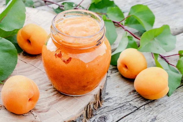 Ahşap bir yüzey üzerinde olgun kayısı ve yeşil yapraklarıyanında cam bir kavanozda kayısı reçeli.