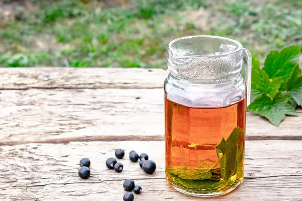 Te i ett glas tekanna med gröna blad av svarta vinbär på ett träbord mot bakgrund av grönt gräs. — Stockfoto