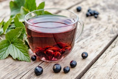 Yeşil yaprakları ve kuş üzümü çilek yanında ahşap bir masada siyah kuş üzümü ile bir fincan çay.