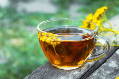Doğanın bir arka plan üzerinde ahşap bir masanın yüzeyinde bir cam kupa ve sarı tansy çiçekler tabak ile bitkisel çay. Tansy Bitkisel çay. Şifa otlar.