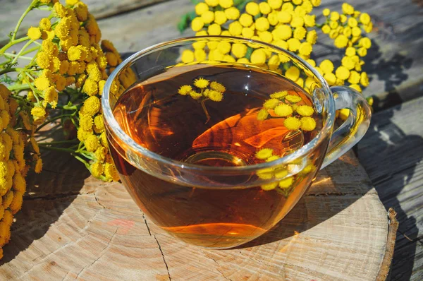 Infuso di tanaceto in una tazza di vetro e fiori di tanaceto gialli su un tavolo di legno. Tansy tisana alle erbe. Erbe curative . — Foto Stock