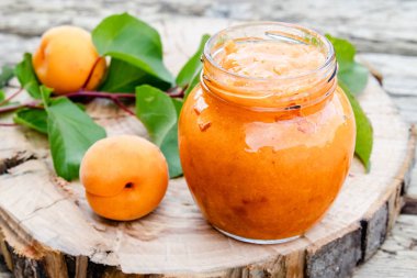 Ahşap bir yüzey üzerinde olgun kayısı ve yeşil yapraklarıyanında cam bir kavanozda kayısı reçeli.