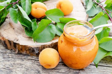 Ahşap bir yüzey üzerinde olgun kayısı ve yeşil yapraklarıyanında cam bir kavanozda kayısı reçeli.