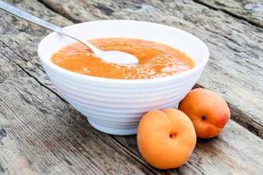 Beyaz bir kapta kayısı reçeli ve tahta bir masada olgun kayısı.