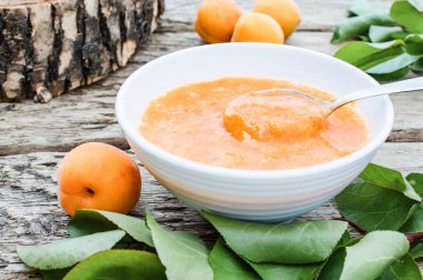 Ahşap bir yüzey üzerinde olgun kayısı ve yeşil yaprakları yanında beyaz bir kapta kayısı reçeli.