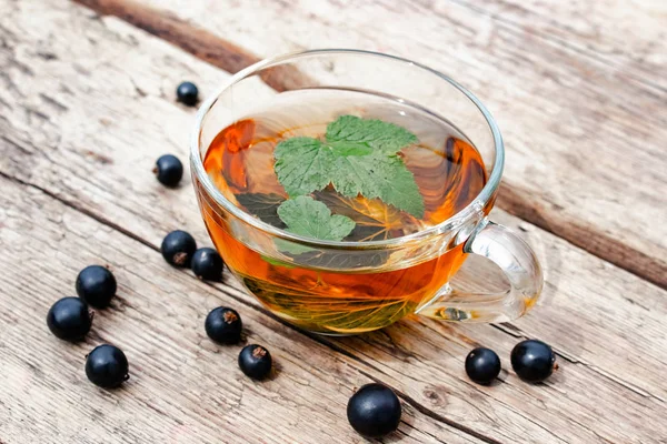 Herbal tea from currant leaves in a glass transparent cup on a wooden table near the green leaves and currant berries.