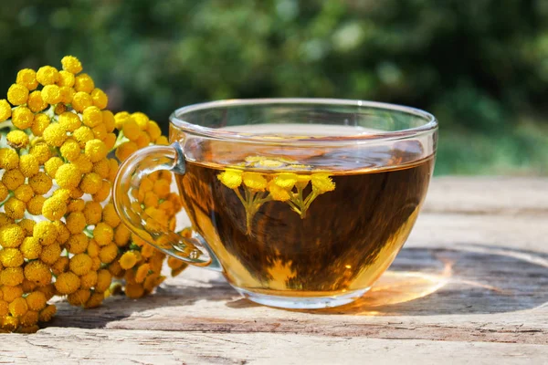 Herbal tea or tansy tincture in a mug and yellow tansy flowers on the surface of a wooden table against the background of nature. Healing herbs. — Stock Photo, Image