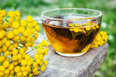 Doğanın arka planına karşı ahşap bir masanın yüzeyinde bir kupa ve sarı tansy çiçekler bitkisel çay veya tansi tentür. Şifa otlar.