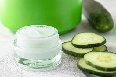 Beyaz ahşap arka planda taze salatalık dilimleri yanında bir cam kavanoz yüz kremi.