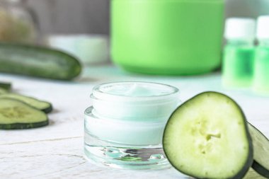 Beyaz ahşap arka planda taze salatalık dilimleri yanında bir cam kavanoz yüz kremi.