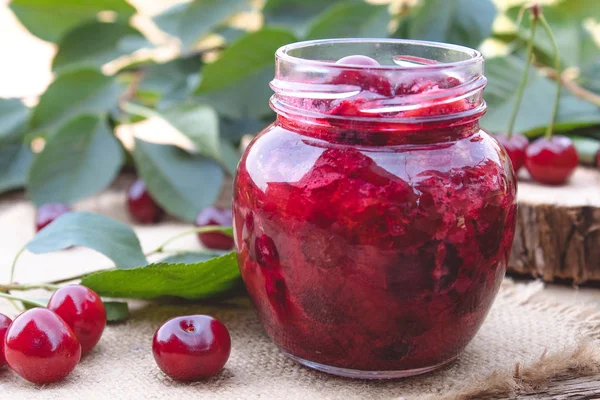 Ahşap tahta bir masada yeşil yaprakları bir arka planda bir cam kavanoz kiraz reçel.