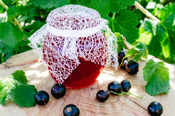 Black currant jam in a glass jar on a wooden table against the currant bush in the garden. Jam from currants. Close-up.