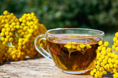 Doğanın arka planına karşı ahşap bir masanın yüzeyinde bir kupa ve sarı tansy çiçekler bitkisel çay veya tansi tentür. Şifa otlar.