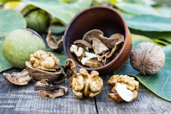 Nueces se encuentran en un tazón junto a nueces en cáscaras verdes y hojas verdes en una mesa de madera vieja rústica . —  Fotos de Stock