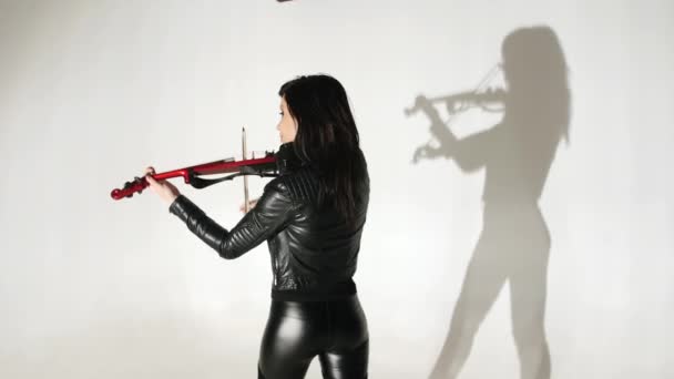 身着紧身黑色皮衣的亚洲少女在白色墙壁上演奏电动小提琴 阴影重复着她的动作 — 图库视频影像
