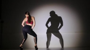 Genç ve güzel bir kadın yüksek topuklularla modern dans ediyor, striptiz dansı, moda, gölge hareketlerini tekrarlıyor.
