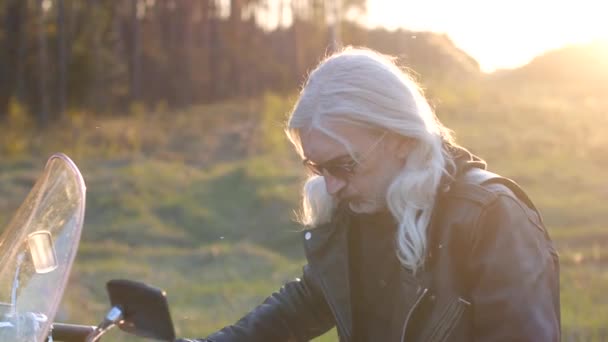 長い灰色の髪を持つ大人の男性の肖像画は 日没時にオートバイに座っています — ストック動画