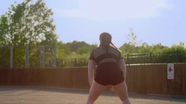 Beyaz tişörtlü, siyah şortlu, sokak dansı yapan, kıvırtan, kalçalarını evlerin ve yeşil ağaçların arkasına doğru sallayan orta boylu, güzel bir kadın dansçı.