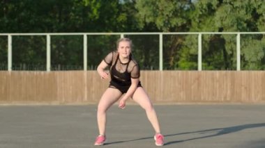 Ağ tişörtlü ve siyah şortlu genç bir kız modern sokak dansında dans ediyor, stadyumda evlerin ve yeşil ağaçların arkasında twerk yapıyor. Gençlik alt kültürü. Çağdaş koreografi. Yavaş çekim