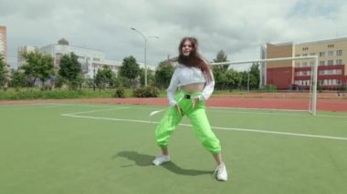Uzun saçlı, eşofmanlı genç bir kadın sokak dansı yapıyor, dans salonu twerk 'i yapıyor, kalçalarını bir spor sahasında sallıyor. Şehirde serbest dans yapan bir şehir. Seks Dansı. Gimbal atışı.